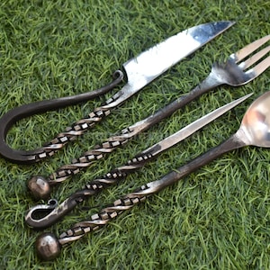 Peut inclure: Ensemble de couverts forgés à la main comprenant un couteau, une fourchette, une cuillère et un couteau à beurre. Les manches ont un design torsadé, et les lames et la cuillère sont argentées. L'ensemble est présenté sur de l'herbe verte.