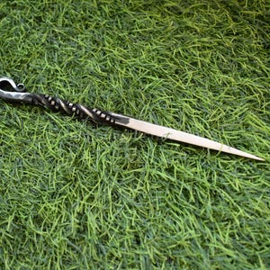 May include: A handcrafted hair stick with a decorative, twisted metal design. The hair accessory features a curved, hook-like top and a pointed end. The stick is set against a backdrop of green grass.