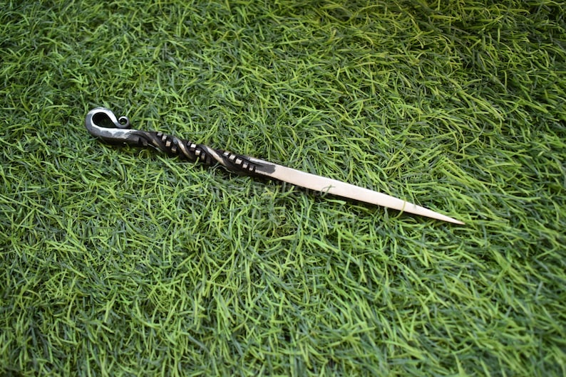 May include: A handcrafted hair stick with a decorative, twisted metal design. The hair accessory features a curved, hook-like top and a pointed end. The stick is set against a backdrop of green grass.