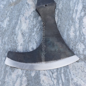 May include: A black metal axe with a curved blade and a hole in the top of the handle. The axe is on a gray and white marble surface. The text "antiquegiftcraftsco" is visible on the blade.