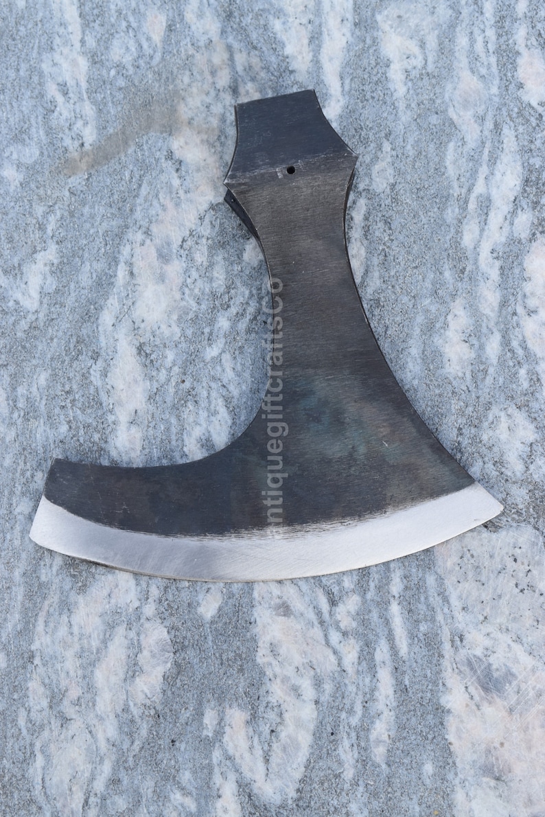 May include: A black metal axe with a curved blade and a hole in the top of the handle. The axe is on a gray and white marble surface. The text "antiquegiftcraftsco" is visible on the blade.