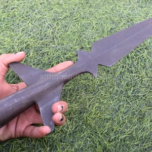 K&ouml;nnte beinhalten: Ein dunkelgrauer Speerkopf mit einer langen, spitzen Klinge und einem stilisierten, fl&uuml;gelartigen Sockel. Der Speerkopf wird vor einem Hintergrund aus gr&uuml;nem Gras gehalten. Das Wort "antiquegifts" ist teilweise sichtbar.
