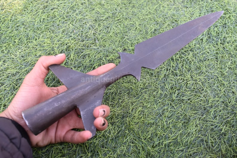 K&ouml;nnte beinhalten: Ein dunkelgrauer Speerkopf mit einer langen, spitzen Klinge und einem stilisierten, fl&uuml;gelartigen Sockel. Der Speerkopf wird vor einem Hintergrund aus gr&uuml;nem Gras gehalten. Das Wort "antiquegifts" ist teilweise sichtbar.