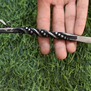 May include: Hand-forged metal skewer with a twisted handle and a curved hook end. The skewer is silver and black, with a textured design. The skewer is held against a green background.