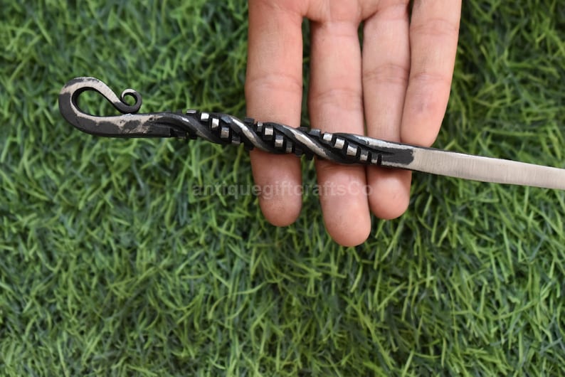 May include: Hand-forged metal skewer with a twisted handle and a curved hook end. The skewer is silver and black, with a textured design. The skewer is held against a green background.