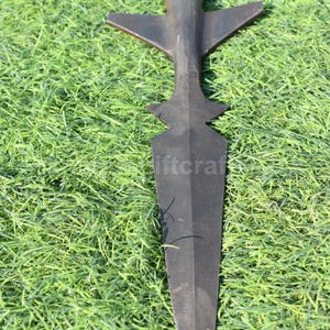 K&ouml;nnte beinhalten: Ein schwarzer Speer mit einer langen, spitz zulaufenden Klinge und einem kreuzf&ouml;rmigen Griff. Das Metallobjekt liegt auf gr&uuml;nem Gras. Die Waffe scheint aus Metall zu sein.