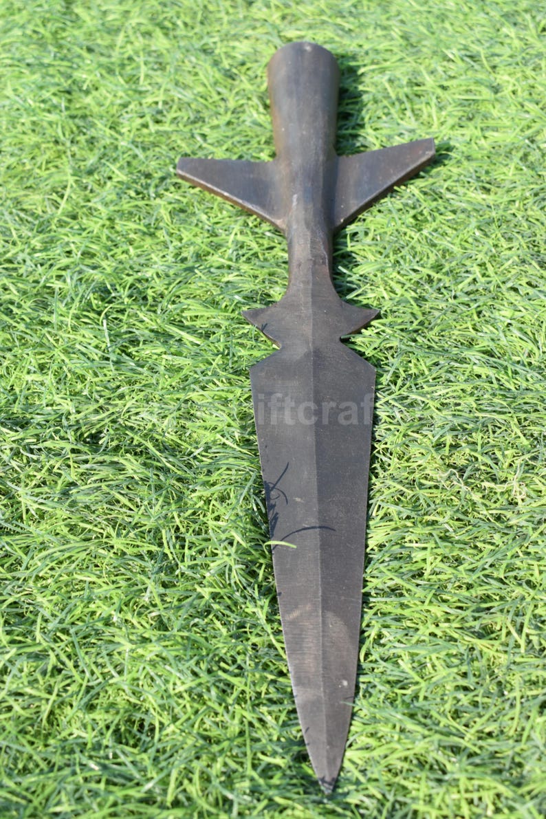 K&ouml;nnte beinhalten: Ein schwarzer Speer mit einer langen, spitz zulaufenden Klinge und einem kreuzf&ouml;rmigen Griff. Das Metallobjekt liegt auf gr&uuml;nem Gras. Die Waffe scheint aus Metall zu sein.
