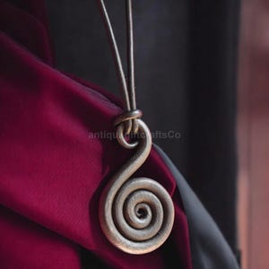 May include: A silver metal pendant with a spiral design hangs from a leather cord. The pendant is on a burgundy fabric background.