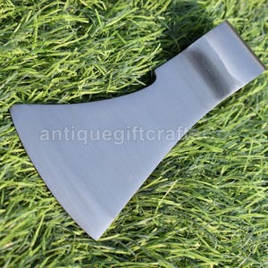 Puede incluir: Una cabeza de hacha de acero con una hoja curva, apoyada sobre hierba verde. La cabeza del hacha tiene el texto "antiquegiftcraft" impreso. La cabeza del hacha es de color gris oscuro.