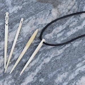 May include: Four bone needles with a black leather cord. The needles are all different lengths and have a pointed tip. The cord is tied to one of the needles.