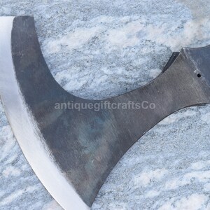May include: A close-up of a hand-forged axe with a curved blade and a dark gray finish. The axe has a wooden handle and a metal head. The axe is on a gray and white marble surface. The text "antiquegiftcraftsCo" is visible on the axe.