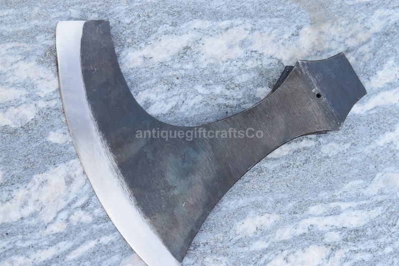 May include: A close-up of a hand-forged axe with a curved blade and a dark gray finish. The axe has a wooden handle and a metal head. The axe is on a gray and white marble surface. The text "antiquegiftcraftsCo" is visible on the axe.