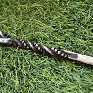 May include: Hand-forged metal fire poker with a twisted shaft and a decorative curled handle. The metal has a dark, textured finish with lighter highlights. The poker rests on a bed of green artificial grass.