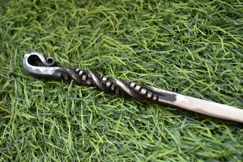 May include: Hand-forged metal fire poker with a twisted shaft and a decorative curled handle. The metal has a dark, textured finish with lighter highlights. The poker rests on a bed of green artificial grass.