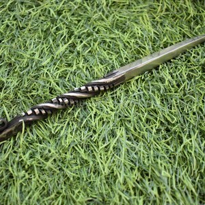 May include: A hand-forged letter opener with a twisted handle and a sharp, pointed blade. The handle is dark, while the blade is silver. The letter opener is lying on a bed of green grass.