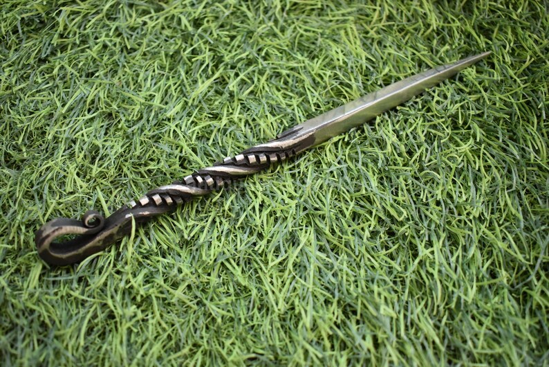May include: A hand-forged letter opener with a twisted handle and a sharp, pointed blade. The handle is dark, while the blade is silver. The letter opener is lying on a bed of green grass.