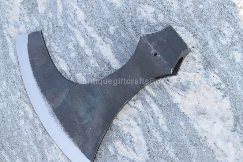 May include: A black metal axe with a curved blade and a handle with a hole in the top. The axe is on a gray and white marble surface.