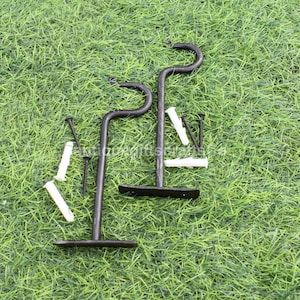 Op de afbeelding: Twee zwarte metalen haken met een gebogen ontwerp, getoond op een groene grasmat. De haken worden geleverd met witte kunststof pluggen en zwarte schroeven.