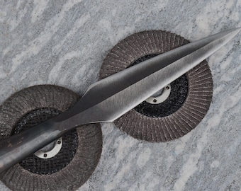 Peut inclure: Gros plan d'une pointe de lance avec une pointe acérée. La pointe de lance en métal est gris foncé et repose sur deux disques de meulage circulaires et texturés. Le fond est une surface grise et blanche mouchetée.