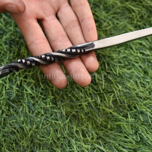 May include: Hand-forged letter opener with a twisted handle and a sharp, silver-colored blade. The handle features a decorative spiral design and a looped end. The opener is held against a backdrop of green grass.