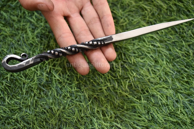 May include: Hand-forged letter opener with a twisted handle and a sharp, silver-colored blade. The handle features a decorative spiral design and a looped end. The opener is held against a backdrop of green grass.