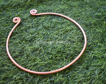 May include: A copper-colored, open-ended necklace with spiral ends, resting on a bed of green artificial grass. The necklace is a simple, circular design, with the text "antiquegiftcraftsCo" visible in the background.
