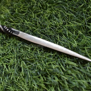 May include: A silver-colored metal hair pin with a twisted handle and a pointed end. The pin is lying on a bed of green artificial grass. The handle has a decorative design.