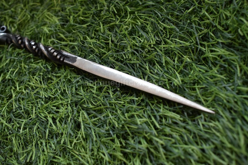 May include: A silver-colored metal hair pin with a twisted handle and a pointed end. The pin is lying on a bed of green artificial grass. The handle has a decorative design.