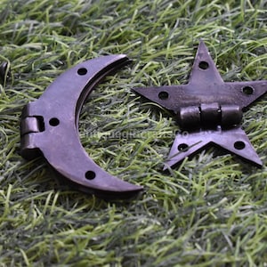 May include: A set of decorative metal hinges in the shape of a crescent moon and a star. The hinges are dark gray with visible screw holes and are placed on a bed of green grass. Several black screws are also visible.