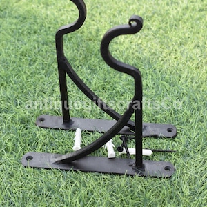 Puede incluir: Soporte de pared de metal negro con dos ganchos curvos. El soporte está fijado a una superficie de hierba verde con anclajes de plástico blanco.