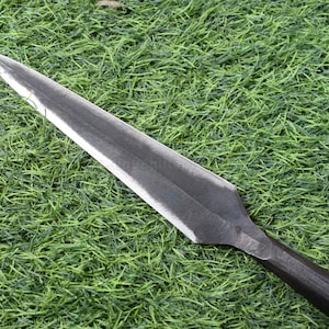 May include: A close-up of a spearhead with a dark, metallic blade and a wooden handle. The spearhead is lying on a bed of green grass. The blade has a sharp point.