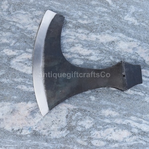 May include: A black metal axe with a curved blade and a wooden handle. The axe is on a gray and white marble surface. antiquegiftcraftsCo