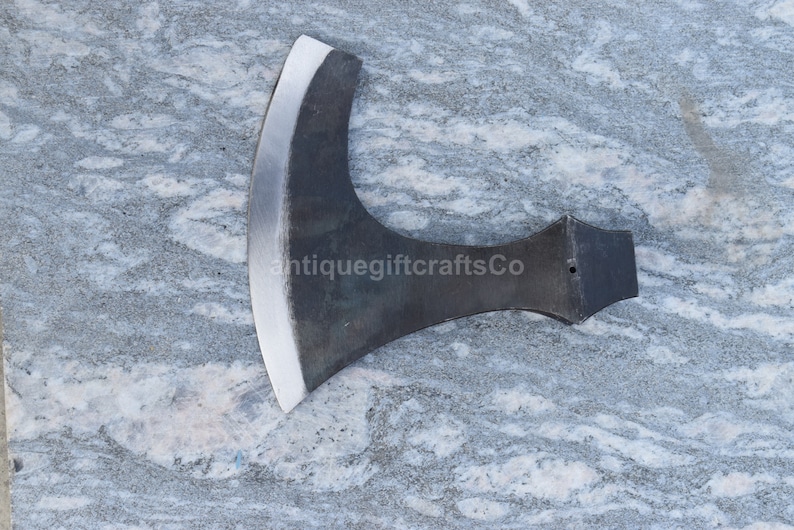 May include: A black metal axe with a curved blade and a wooden handle. The axe is on a gray and white marble surface. antiquegiftcraftsCo