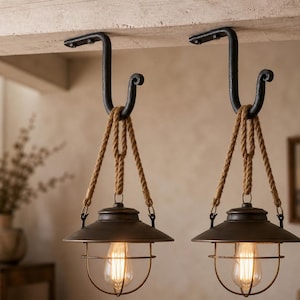 Może przedstawiać: Dwie wiszące lampy o rustykalnym, industrialnym designie. Każda lampa ma ciemny metalowy klosz, przezroczystą szklaną obudowę i żarówkę Edison w stylu vintage. Światła są zawieszone na grubych, plecionych linach z czarnych metalowych haczyków.