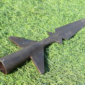K&ouml;nnte beinhalten: Ein dunkler, antiker Speerkopf mit einer langen, spitzen Klinge und einem zylindrischen Sockel. Der Speerkopf hat zwei Seitenfl&uuml;gel und liegt auf einem Bett aus gr&uuml;nem Gras. Der Speerkopf ist aus Metall.