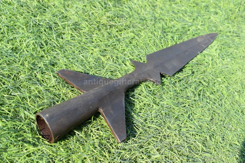 K&ouml;nnte beinhalten: Ein dunkler, antiker Speerkopf mit einer langen, spitzen Klinge und einem zylindrischen Sockel. Der Speerkopf hat zwei Seitenfl&uuml;gel und liegt auf einem Bett aus gr&uuml;nem Gras. Der Speerkopf ist aus Metall.