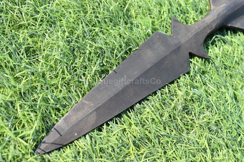 K&ouml;nnte beinhalten: Ein schwarzer Speerkopf mit einer Spitze und einem sternf&ouml;rmigen Design in der N&auml;he des Griffs. Der Speerkopf liegt auf einem Bett aus gr&uuml;nem Gras. Der Text "uegiftcraftsCo" ist sichtbar.