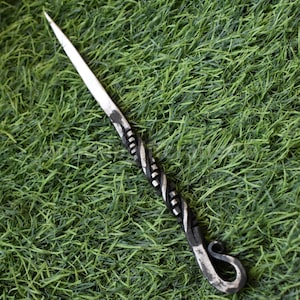 May include: A hand-forged metal awl with a sharp, pointed tip and a decorative, twisted handle. The handle transitions from a dark, textured finish to a silver-toned, polished metal. The awl has a looped end. The awl is set against a green background.