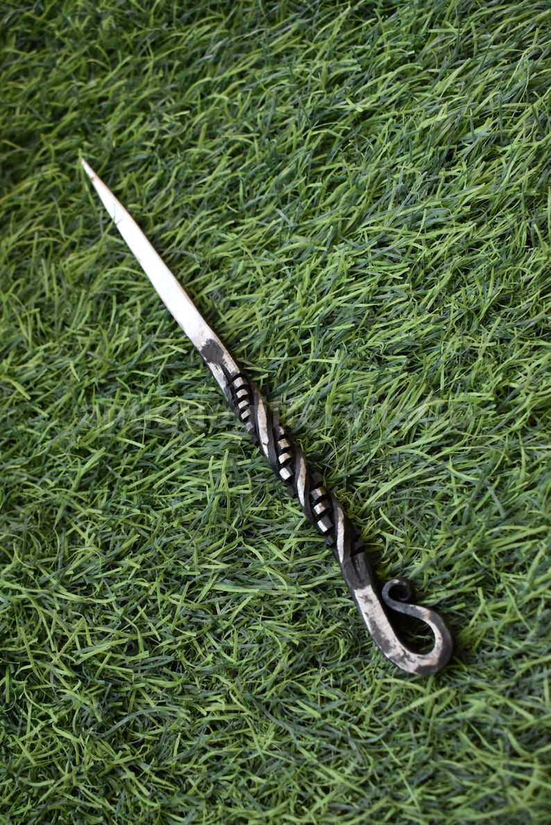 May include: A hand-forged metal awl with a sharp, pointed tip and a decorative, twisted handle. The handle transitions from a dark, textured finish to a silver-toned, polished metal. The awl has a looped end. The awl is set against a green background.