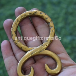 May include: A golden-coloured metal hook with a hammered texture. The hook has a circular top and a curved, stylized bottom. The text "antiquegiftcraftsCo" is visible on the hook. The hook is likely a decorative item.