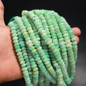 Può includere: Una collana di perle di opale verde, ogni perla è rotonda e liscia. Le perle sono disposte in una pila, mostrando la variazione di colore e texture.
