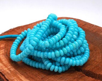 May include: A strand of turquoise blue beads, each bead is round and slightly elongated, creating a textured look. The beads are piled on a wooden surface.