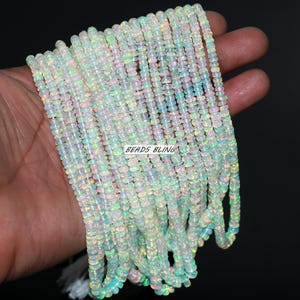 Puede incluir: Hebras de cuentas de ópalo iridiscentes. Las cuentas son pequeñas, planas y redondas, con una mezcla de colores blanco, azul y verde. El texto "BEADS BLING" es visible en la imagen. Las cuentas están ensartadas, creando una apariencia fluida y texturizada.