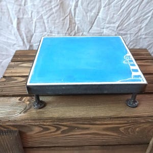 Puede incluir: Una baldosa de cerámica azul y blanca con un diseño de faro, montada en un soporte de metal negro con cuatro patas. El soporte está sentado sobre una superficie de madera.