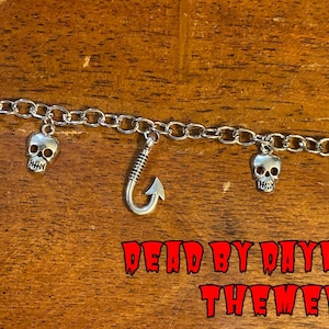 Pode incluir: Uma pulseira de corrente prateada com quatro berloques de caveira e um berloque central de anzol. A pulseira inclui um fecho de barra. O texto "DEAD BY DAYLIGHT THEMED" está impresso em vermelho e um logotipo gráfico branco é visível.