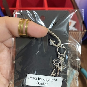 May include: A silver-toned metal charm featuring a fishhook, a stethoscope, and a lightning bolt shape attached to a black display card. The card has a white label with the black text Dead by daylight Doctor printed on the front surface.