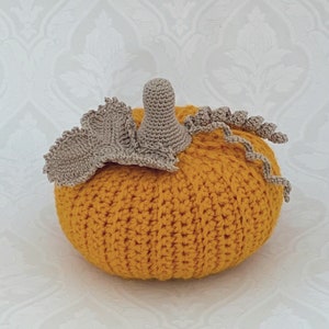 May include: A crocheted pumpkin with a stem and leaf detail. The pumpkin is a bright yellow color and the stem and leaf are a light brown color.