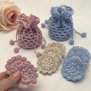 May include: Three crocheted drawstring bags in pink, blue, and beige. Each bag has a scalloped top and is decorated with small pom-poms. The bags are arranged on a white surface.