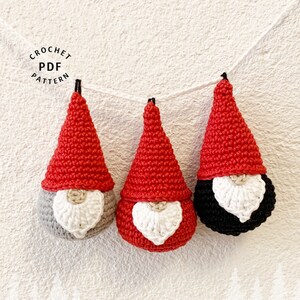 May include: Three crocheted gnomes with red hats, white beards, and different colored bodies: gray, red, and black. The gnomes are hanging from a string.