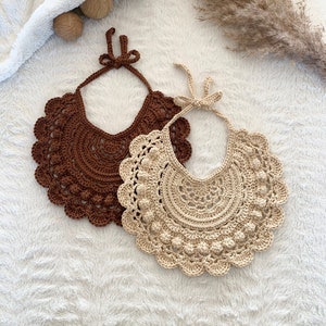 May include: Two crocheted bibs, one brown and one beige, with a scalloped edge and a center circle of raised stitches. The bibs are laid out on a white fluffy surface.
